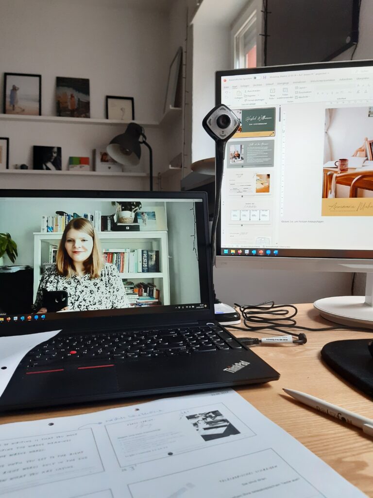 Zwei Bildschirme, mehrere Kameras, eine Powerpoint-Präsentation und Arbeitsblätter auf einem Schreibtisch